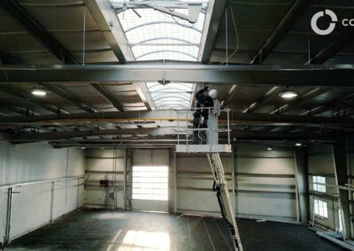 Pracovník na rebríku vykonáva údržbu v priemyselnej hale.